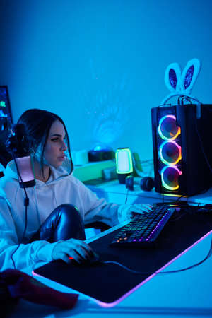 Happy woman putting on headphones and playing computer game. Female gamer in glasses sitting and...