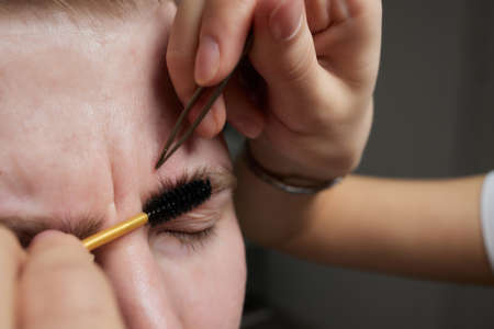 Professional barber doing threading procedure and correcting shape of eyebrows to young male client...