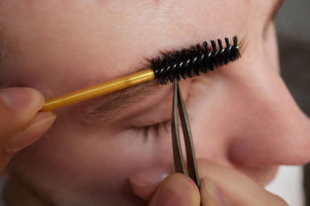 Professional barber doing threading procedure and correcting shape of eyebrows to young male client...