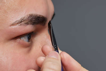 Professional barber doing threading procedure and correcting shape of eyebrows to young male client with tweezer in barber shop. Barber at work. Brow care concept. Plucking brows.の写真素材