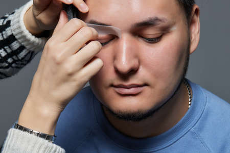 Professional barber doing threading procedure and correcting shape of eyebrows to young male client...
