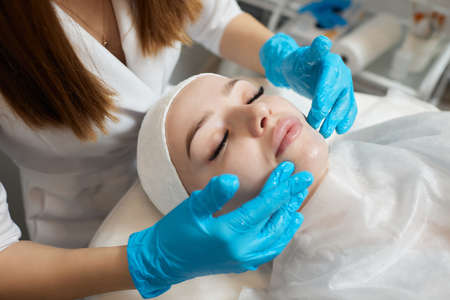 Mirthful young woman lying in a shower cap and hands in rubber gloves cleaning her skin with cotton...