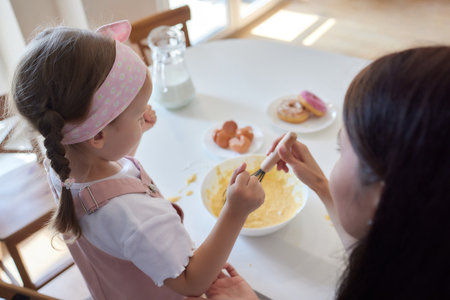 Mother and daughter preparing a sweet cake using flour, milk, sitting on chairs at a table in a...