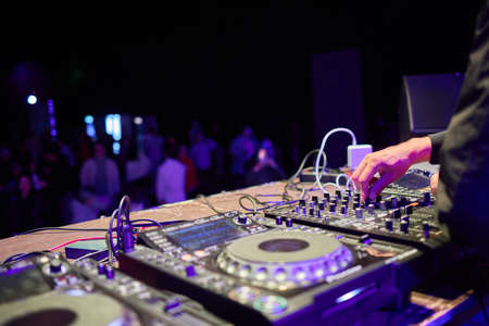 control DJ for mixing music with blurred people dancing at party in nightclub.
