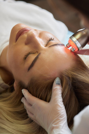 A cosmetologist in rubber gloves does the rf-lifting procedure on the face. In the background, the Rf cosmetology device. Close up. Side view. The concept of professional cosmetology and rejuvenation.の写真素材