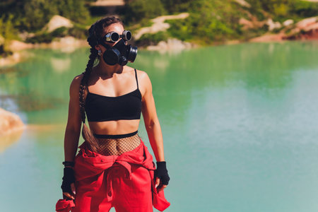 Woman dressed in a steampunk suit in a defiant attitude with her arms crossed. respirator on the face, against the background of a dirty lakeの写真素材