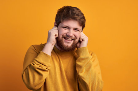 Portrait of attractive unshaven male pluggs ears as hears annoying sound, frowns face, clenches teeth, models indoor. Hipster doesn t want to hear noise ignores something unpleasant, can t stand crash.の写真素材