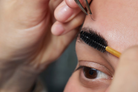 Professional barber doing threading procedure and correcting shape of eyebrows to young male client with tweezer in barber shop. Barber at work. Brow care concept. Plucking brows.の写真素材
