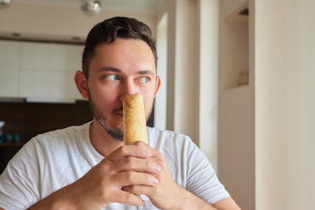 Closeup portrait of hungry man eating delicious sandwich at home on background of kitchen. Handsome...