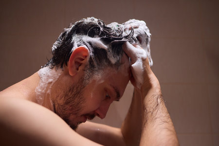 Man washing hair with shampoo in shower, creating foamy lather - personal hygiene care.