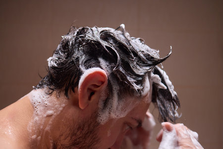 Man washing hair with shampoo in shower, creating foamy lather - personal hygiene care.