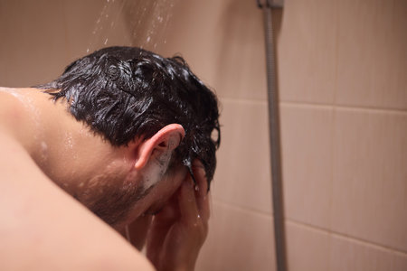 Man washing hair with shampoo in shower, creating foamy lather - personal hygiene care.