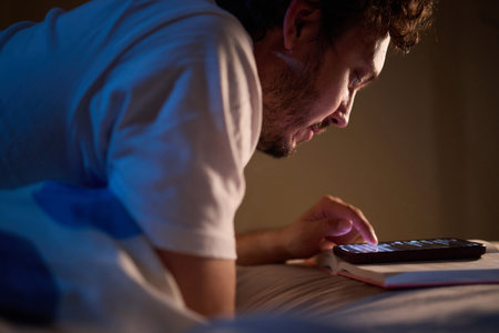 A man is peacefully reading a book in bed in a dimly lit bedroom, covered with cozy blanketsの写真素材