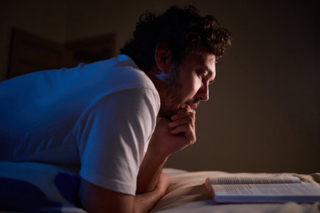 A man is peacefully reading a book in bed in a dimly lit bedroom, covered with cozy blanketsの写真素材