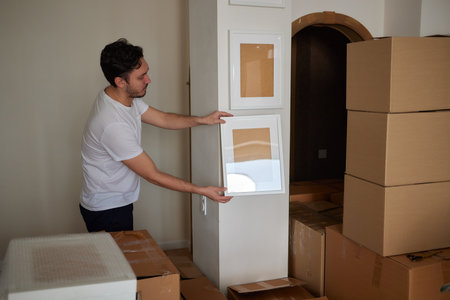 A young man crouched down looking through a moving box, filled with clothes, books and a picture frame.の写真素材