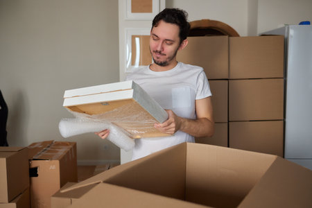 A young man crouched down looking through a moving box, filled with clothes, books and a picture frame.の写真素材