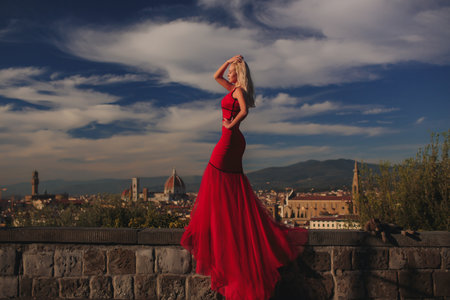 A Stunningly Elegant Woman Dressed in a Red Dress Against a Beautiful Scenic Cityscapeの写真素材