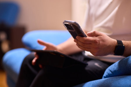 A person is seen sitting comfortably on a blue couch while using both a smartphone and a tabletの写真素材