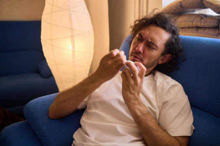 A man is sitting on a blue couch, holding a tissue and blowing his nose. He looks uncomfortable and is probably feeling unwell.の写真素材