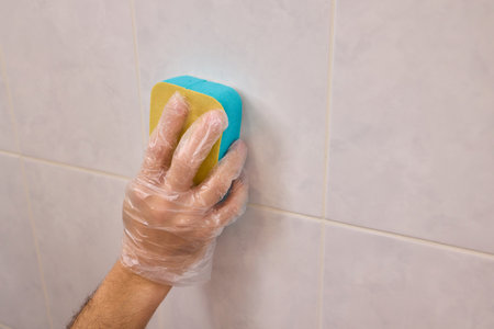 A Man Diligently Cleaning a Tiled Bathroom Wall with His Gloves and a Scrubber Tool