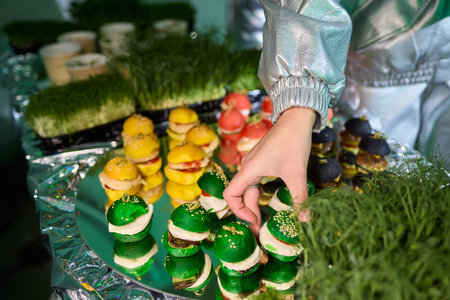 Colorful miniature burger-shaped macarons displayed on shiny green foil surface, Playfully arranged...