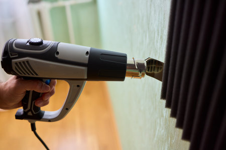 Craftsman carefully detaching acoustic panels indoors, Individual skillfully peels acoustic insulation from wall using heat and precise hand movements for studio setupの写真素材
