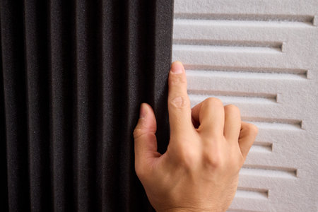 Closeup of foam insulation method, Hand pressing textured acoustic foam onto wall surface, Detailed shot of fingers securing black foam panels on gray wall Comparison of different foam rubber and sound absorption efficiencyの写真素材
