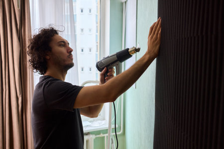 Craftsman carefully detaching acoustic panels indoors, Individual skillfully peels acoustic insulation from wall using heat and precise hand movements for studio setupの写真素材