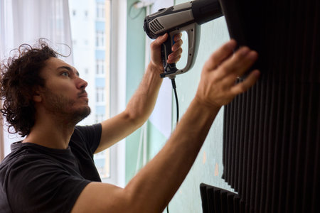 Craftsman carefully detaching acoustic panels indoors, Individual skillfully peels acoustic insulation from wall using heat and precise hand movements for studio setupの写真素材