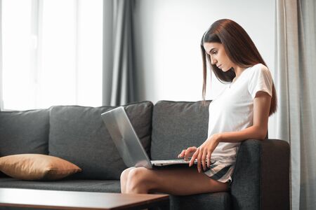 Young woman with modern laptop sitting on sofa at home. Happy girl browsing through the internet during free time.. の写真素材
