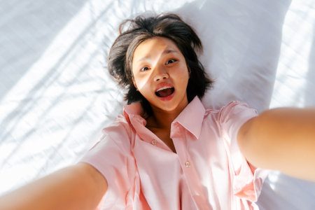 Asian young female lying on bed smiling up at camera in pink shirt.の写真素材