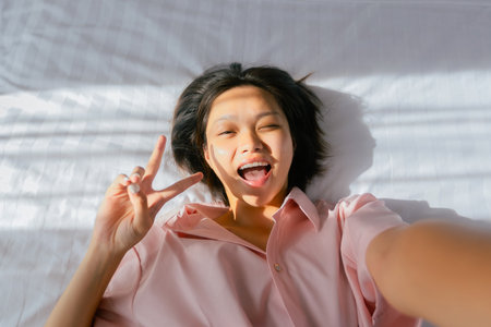 Smiling asian young female making peace sign in sunlit room.の写真素材