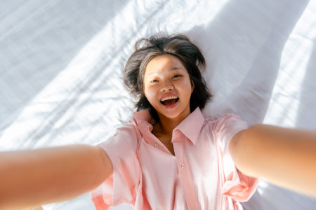 Joyful asian female teen lying on bed in pink shirt with sunlight shadows.の写真素材