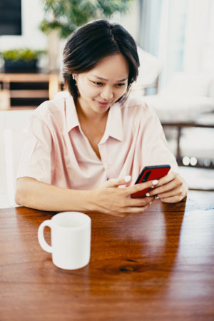 Asian young adult female using smartphone in modern home interior with coffee mug.の写真素材