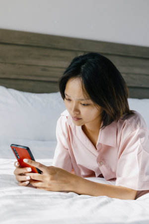 Asian young woman relaxing on bed using smartphone in casual pink shirt.の写真素材