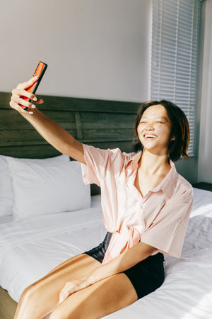 Asian young female taking selfie in bedroom with natural light.の写真素材