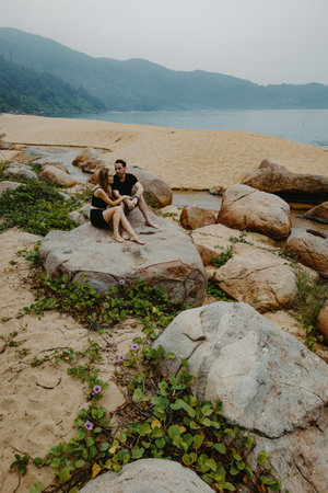 Young caucasian couple relaxing on rocky beach with scenic coastal view.の写真素材