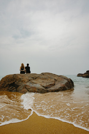Serene beach scene with two young adults relaxing on rock by ocean shoreline.の写真素材