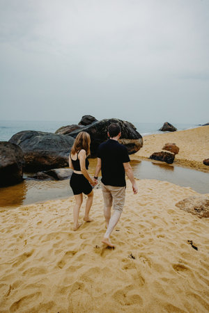 Young adult caucasian couple walking on sandy beach by rocks.の写真素材