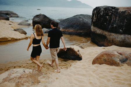 Young caucasian couple walking on a rocky beach hand in hand at sunset.の写真素材