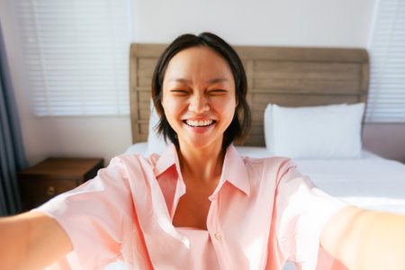 Smiling asian female taking selfie in bright bedroom.の写真素材