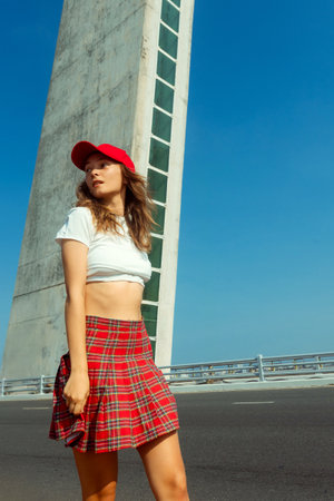 Young caucasian female in red cap and plaid skirt walking near tall building.の写真素材