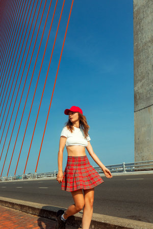 Young caucasian female in red plaid skirt walking on bridge under blue sky.