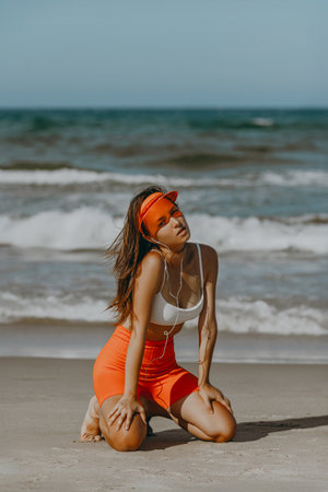 Young caucasian female in sporty attire kneeling on beach with headphones.の写真素材