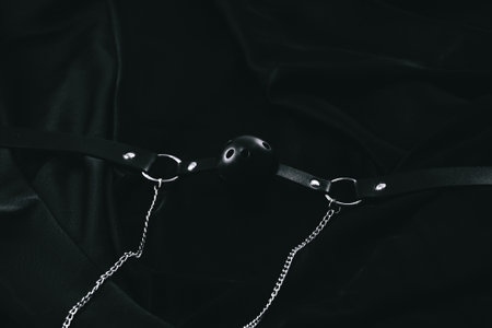 Black leather ball gag with metal chains on dark fabric background.の写真素材