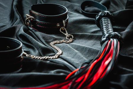 Black leather restraints and red-tipped flogger on dark fabric background.