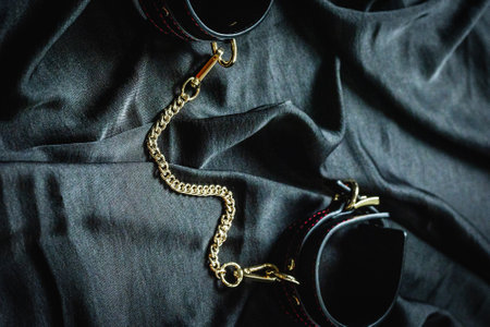 Black leather handcuffs with gold chain on dark fabric surface.