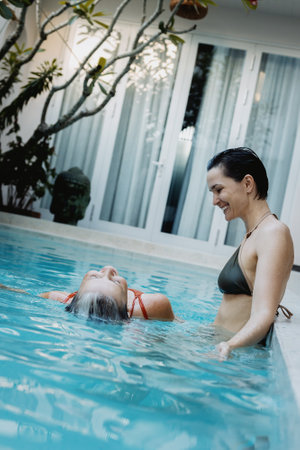 Asian female adult friends enjoying relaxing swim in backyard pool retreat.