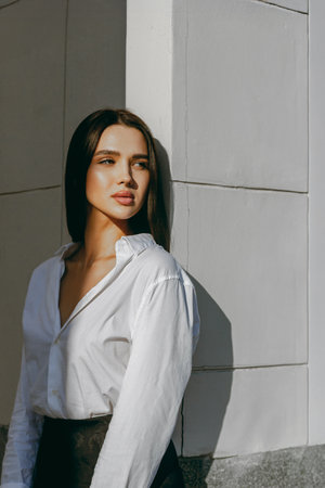 Caucasian young female in white shirt leaning against a wall in bright sunlight.の写真素材