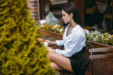 Young adult caucasian female drinking coffee outdoors by flowers.の写真素材